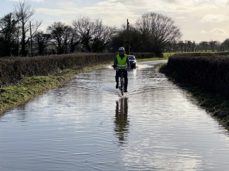 Senior man and car driving through a flooded country lane in Nunney, Somerset after heavy rain. February 2025
